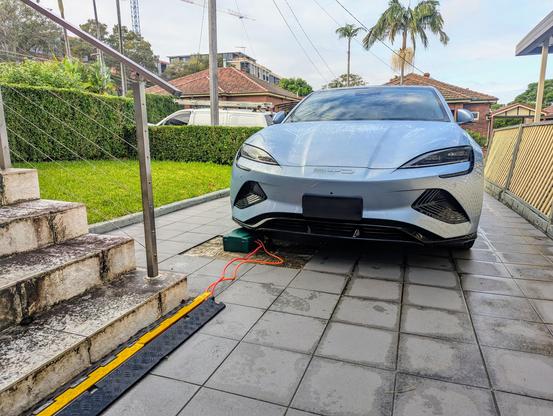 Photo of a parked BYD Seal electric car from the front. There is a green box on the ground by the front of the car and two cables running out of that box towards and past the photographer.