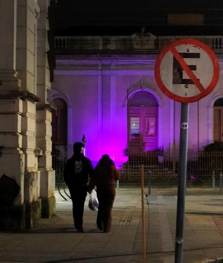 A imagem mostra duas pessoas caminhando de costas para a câmera em uma rua à noite. Elas estão próximas umas das outras, com uma das pessoas segurando uma sacola plástica. A iluminação é suave, com uma luz roxa projetada na fachada de um edifício clássico ao fundo, que possui arcos e janelas. Um sinal de trânsito proibindo estacionamento, com a letra "E" dentro de um círculo vermelho e uma linha diagonal, está visível à direita. A cena transmite uma atmosfera tranquila e urbana, com a iluminação noturna e a arquitetura clássica do edifício.