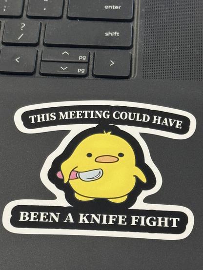 A laptop palm rest area showing keys above. There is a sticker on the palm rest showing a cute cartoon duck holding a knife, with the caption, “This meeting could have been a knife fight.”