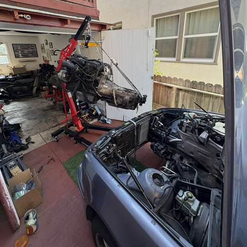 The M54B25 inline six-cylinder engine and ZF 5HP19 five-speed automatic transmission hang from a hoist in front of the vacant engine compartment of my blue 2003 BMW 5-series station wagon.