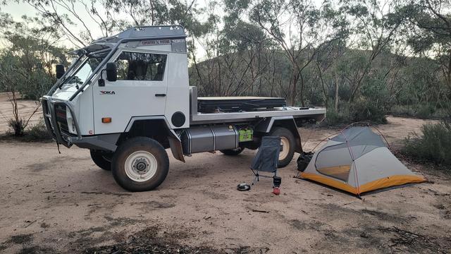 A small tent, chair and camp stove next to an OKA truck parked on some sandy soil amongst trees