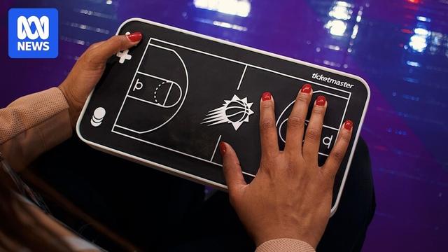 A black, flat model basketball field is being held by one hand and another hand is on its surface with bright red nail polish.