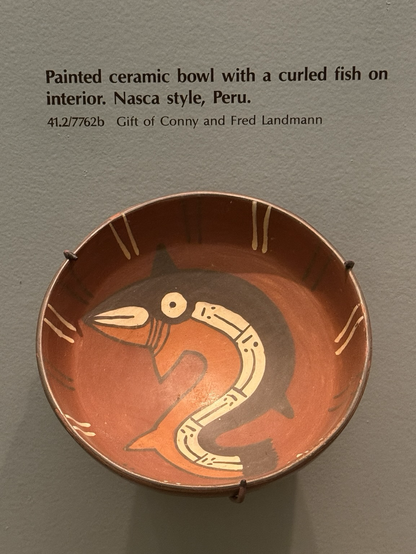 photo of polychrome ceramic bowl on display at museum with gallery label: “Painted ceramic bowl with a curled fish on interior. Nasca style, Peru. 41.2/7762b Gift of Conny and Fred Landmann”