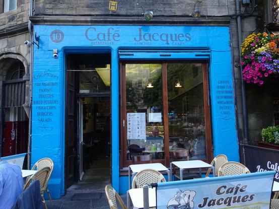 Fachada de un café, pintada de un color azul intenso, en el casco antiguo de una ciudad. Edificio de piedra. Varias mesas con sillas en el exterior. No se ve gente. En el tótulo: "10 - Café Jacques - Breakfast Lunches  Cakes".