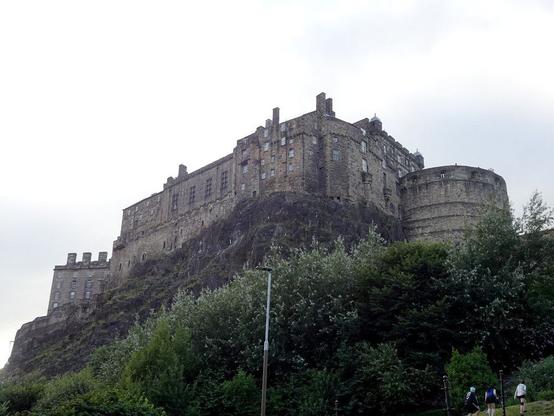 Un castillo de gran tamaño en lo alto de una colina, visto desde abajo. En primer plano, abajo, árboles. En la esquina inferior derecha, tres personas, apenas visibles.