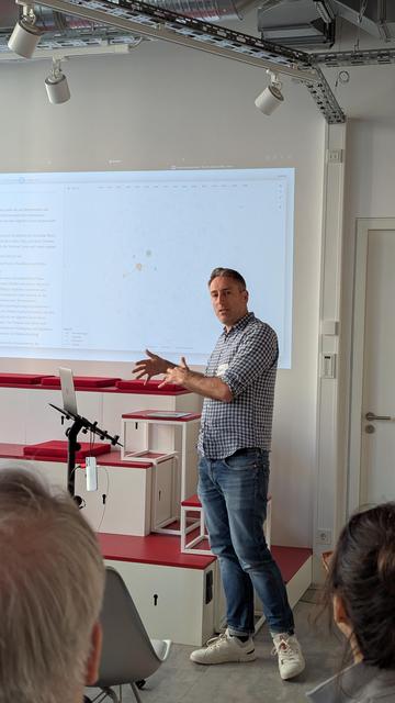 Das Bild zeigt eine Person, die eine Präsentation vor einem Bildschirm hält. Auf dem Bildschirm ist ein Diagramm mit verbundenen Knoten zu sehen, das vermutlich eine Datenvisualisierung oder Netzwerkstruktur darstellt. Die Person gestikuliert mit beiden Händen und erklärt möglicherweise die Inhalte des Diagramms. Der Raum wirkt modern und ist mit Spotlights sowie einem Projektor ausgestattet. Mehrere Zuschauer verfolgen aufmerksam die Präsentation.
