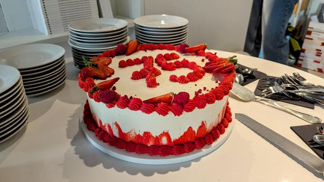 Das Bild zeigt eine dekorierte Torte, die auf einem Tisch steht. Sie ist weiß glasiert und mit roten Verzierungen aus Zuckerguss sowie frischen Erdbeeren und Himbeeren geschmückt. Auf der Oberfläche der Torte steht die Zahl ‚25‘ in roter Glasur geschrieben. Rund um die Torte liegen weiße Teller, silbernes Besteck und Servietten. Im Hintergrund sind einige aufgestapelte Bücher oder Notizhefte sichtbar.