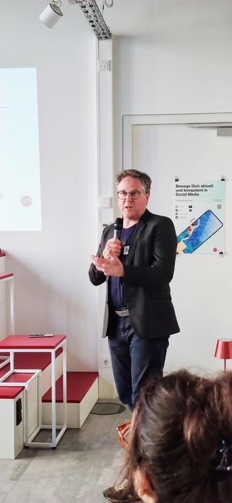 Das Bild zeigt eine Person, die in einem modernen Seminarraum einen Vortrag hält. Die Person trägt einen schwarzen Blazer, ein dunkles Hemd und Jeans und spricht in ein Mikrofon. Im Hintergrund ist eine weiße Wand mit einem Poster, auf dem 'Bewege Dich aktuell und kompetent in Social Media' steht. Das Poster zeigt auch Symbole verschiedener Social-Media-Plattformen und einen QR-Code. Links befindet sich eine Leinwand mit einer Präsentation, die das Wort 'love' und andere, nicht lesbare Inhalte zeigt. Im Vordergrund sind rote und weiße Sitzstufen sowie eine kleine rote Lampe zu sehen.
