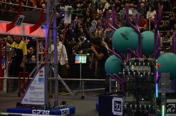 A photo of an FRC robot extending its metal elevator mechanism upward, in preparation to place the piece of PVC pipe it holds on top of the metal structure at right.