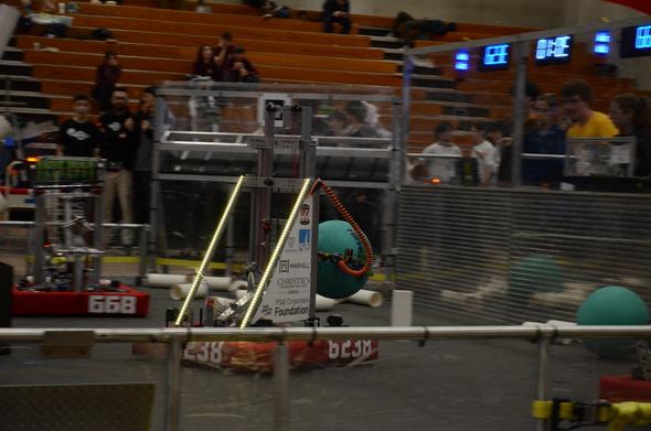 A motion-blurred photo of an FRC robot picking up a large teal playground ball. The robot is clear, but everything around it is blurred.