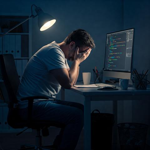 Ein Mann sitzt in einem dunklen Raum im Schein einer Lampe vor einem Computerbildschirm und hat sein Gesicht in den Händen vergraben. Auf dem Computerbildschirm ist Programmcode zu sehen.