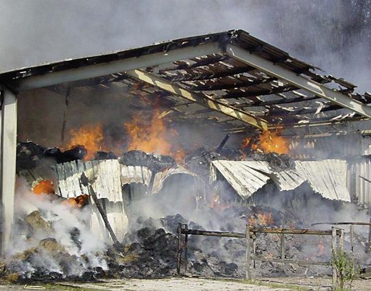Das Bild zeigt einen brennenden Schuppen mit dicken Rauchwolken, während Flammen aus den Wänden und dem Dach schlagen. Teile des Daches sind beschädigt und hinuntergefallen, und der Boden ist mit verbranntem Material bedeckt. Der Hintergrund ist verschwommen und zeigt möglicherweise mehr Rauch und Feuer.