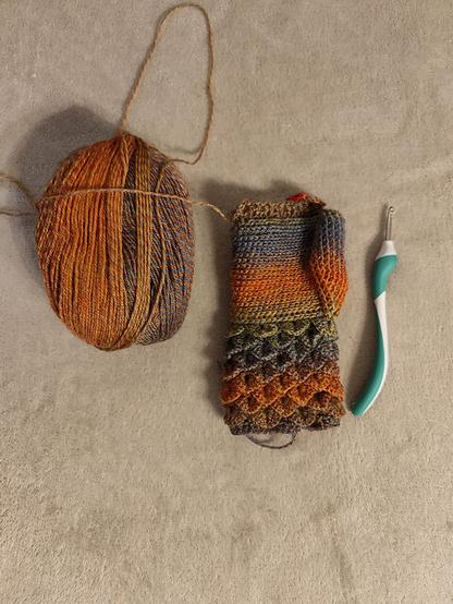 A ball of multicolored yarn in shades of orange, brown, and blue rests on a beige surface alongside a partially crocheted mitt. A white crochet hook with a green grip lies nearby, signifying an ongoing crafting project. The intricate stitch pattern suggests a skilled hand at work, creating a warm and textured accessory.