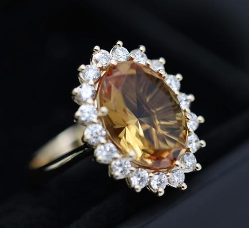 Close-up of a custom-made ring featuring a 3.3ct oval-cut colour-changing sultanite gemstone, surrounded by a halo of 18 round white diamonds, all set in a 9ct yellow gold band, displayed on a black background.