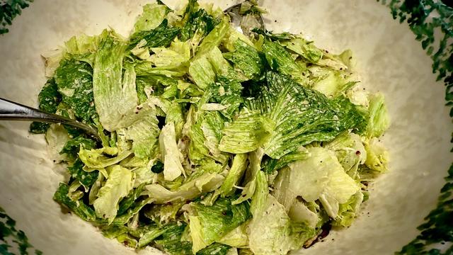 This is an image of Caesar salad in a ceramic bowl with a green rim. The lettuce is torn and dressing and cheese coats the leaves. It looks very tasty.