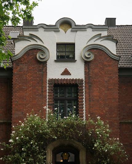 Façade de maison de ville en brique et je ne sais pas trop quoi, stuc et béton peut-être. La partie haute est ornée d‘arabesques et autres éléments décoratifs qui évoquent des sourcils froncés. Il y a de la végétation qui fait comme une moustache au-dessus de la porte d’entrée.