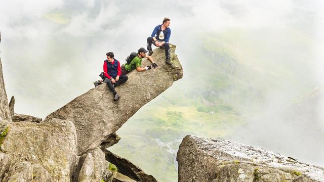 Three male figures, two sitting, one prone, perch on a finger of rock that projects, tilted at a rising angle, several metres out from a rocky ridge. Clouds swirling below reveal enigmatic glimpses of the wider landscape, with a lake, hill slopes, a road, and a small cluster of buildings and parked cars.
