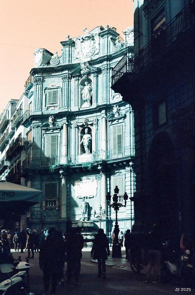 Blick auf eine der vier Ecken des Platzes Quattro Canti in Palermo, Sizilien, Italien, das eine der Barockfassaden mit Skulpturen in den Nischen in der Mitte und hohen Fenstern links und rechts zeigt. Im Vordergrund sehr dunkel Passanten. Verfremdete Farben in Richtung grün und braun wegen des verwendeten Lomochrome Turquoise-Films.