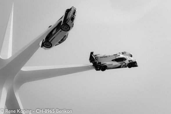 Das Bild zeigt zwei Porsche, die in der Luft schweben und der Schwerkraft zu trotzen scheinen, befestigt an einer großen, weißen, sternförmigen Struktur. Die Struktur hat mehrere Arme, die sich nach außen erstrecken, wobei die Autos an den Enden positioniert sind. Der Wagen auf der linken Seite ist ein schnittiger, moderner Sportwagen mit silberner Karosserie und schwarzen Akzenten, während der Wagen auf der rechten Seite ein Rennwagen mit überwiegend weißer Karosserie und schwarzen Details ist, auf denen die Nummer "2" und verschiedene Sponsorenlogos zu sehen sind. Der Hintergrund ist ein schlichter, hellgrauer Himmel, der die Autos und die Struktur hervorhebt. Das Bild ist in Schwarz-Weiß gehalten, was der Szene einen dramatischen Effekt verleiht. Die Autos sind unterschiedlich geneigt, wobei der Sportwagen nach oben und der Rennwagen leicht nach unten geneigt ist. Die Gesamtkomposition erzeugt ein Gefühl von Bewegung und dynamischer Spannung.
*** Übersetzt mit www.DeepL.com/Translator (kostenlose Version) ***