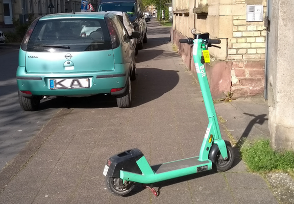 Scooter von Bolt steht quer über Gehweg, vor halb auf Gehweg parkendem Auto und zwingt Fußgänger zum Slalom.