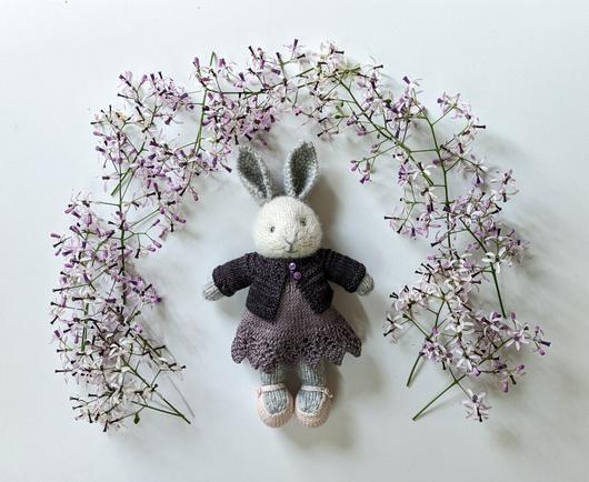 A small, handknitted rabbit wearing a light purple linen dress and a dark purple cardigan.