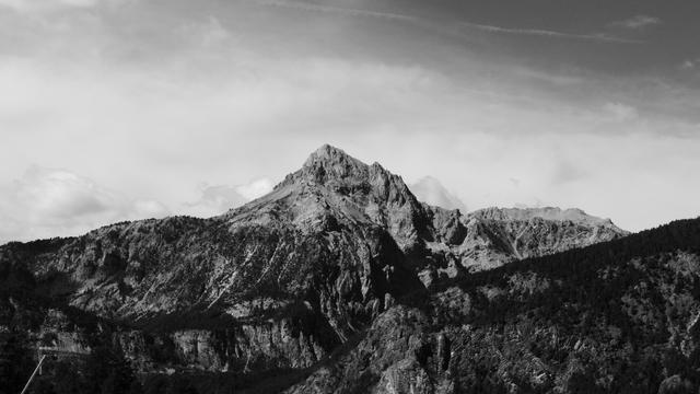 This is a black and white photograph of a mountainous landscape. It features a prominent mountain peak with rugged slopes, surrounded by smaller peaks and ridges. The sky is partly cloudy.