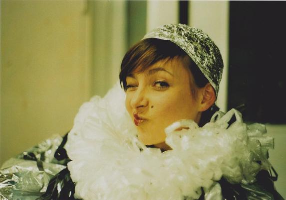 Portrait of a young woman with sleek dark hair, black eyeliner and red lips. Her expression is a cheeky kiss to the side, while she is looking at the camera. She wears a cap made of aluminium foil and a big voluminous collar made of plastic bags. The rest of her outfit (hardly visible) is also of aluminium foil.