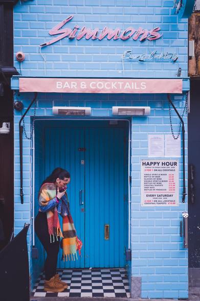 A woman with a colourful scarf stands in front of the entrance to Simmons Bar & Cocktails. The bar's facade is clad in azure tiles, with a pink awning casting a muted glow.
