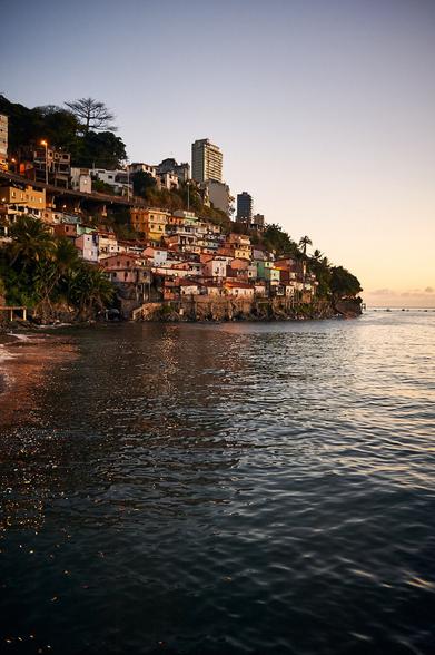 The front part until the middle shows the sea, with bits of a pebble beach to the left side. From the middle a steep coastline with houses built from sea level until the top of the hill are seen. Above a blue sky with the last light of day is displayed.
