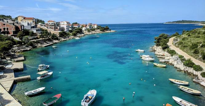 An elevated, wide shot captures a picturesque coastal inlet filled with clear, turquoise water dotted with numerous small boats. The boats, mostly white with some showing colourful accents, are moored in the calm bay. The coastline on either side is rocky and lined with Mediterranean vegetation, primarily green trees and shrubs.
On the left side of the inlet, a cluster of buildings, predominantly white and terracotta-roofed houses, are nestled along the shore and ascend the gentle slope. Stone walls and steps lead down to the water's edge, indicating human habitation and access to the sea.
The right side of the inlet features a more natural, less developed shoreline with a winding dirt path visible through the greenery. In the distance, the deep blue of the open sea stretches out to the horizon under a partly cloudy, light blue sky. A small, green island is visible in the distance, adding to the scenic view.