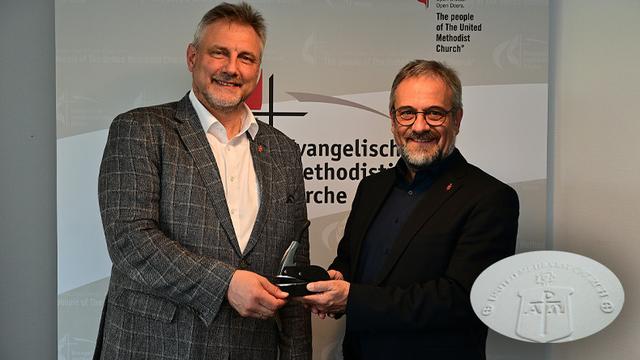 Zwei Männer mittleren Alters stehen sich gegenüber und halten das Dienstsiegel in den Händen. Der Mann links trägt einen grauen, gemusterten Blazer, der Mann rechts einen dunklen Anzug. Beide Männer lächeln und tragen ein rotes Abzeichen an der Kleidung. Im Hintergrund ist ein Banner mit dem Text "The people of The United Methodist Church" zu sehen.