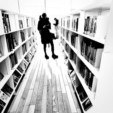 A silhouette of a person standing between bookshelves in a bright, modern library, browsing through books on a wooden floor.