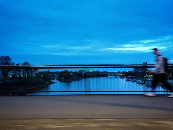 A long exposure shot taken at dusk shows a cityscape bordering a wide river. The sky is a deep blue with some lighter streaks near the horizon, suggesting the fading or approaching light. The river, reflecting the sky and the city lights, flows horizontally across the frame, its surface slightly blurred due to the long exposure.
Along both banks of the river, buildings are illuminated with warm yellow lights, creating a contrast with the cool blue tones of the sky and water. Trees line the riverbanks, their dark silhouettes visible against the city lights. Several boats are docked along the river, their lights adding to the urban glow.
In the foreground, a pedestrian is captured in motion, walking from left to right across what appears to be a bridge. The long exposure has blurred the person's form, indicating movement. The bridge has metal railings that run horizontally across the lower half of the frame, slightly obscuring the view of the river and the opposite bank.