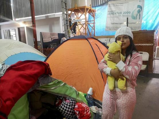 Das Foto zeigt María Orellana mit Sofia auf dem Arm, stehend vor einigen Zelten.