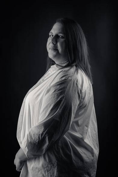 A black-and-white portrait of a woman standing confidently against a dark background. She wears a white button-down shirt over a lace top, with her hands holding the shirt closed. Her long hair falls to one side, and she looks directly at the camera with a calm, self-assured expression. The lighting emphasizes her strong presence and natural beauty.