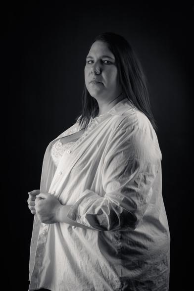 A black-and-white portrait of a woman standing confidently against a dark background. She wears a white button-down shirt over a lace top, with her hands holding the shirt closed. Her long hair falls to one side, and she looks directly at the camera with a calm, self-assured expression. The lighting emphasizes her strong presence and natural beauty.