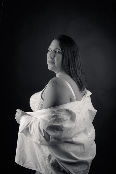A black-and-white portrait of a woman standing confidently against a dark background. She wears a white button-down shirt over a lace top, with her hands holding the shirt closed. Her long hair falls to one side, and she looks directly at the camera with a calm, self-assured expression. The lighting emphasizes her strong presence and natural beauty.