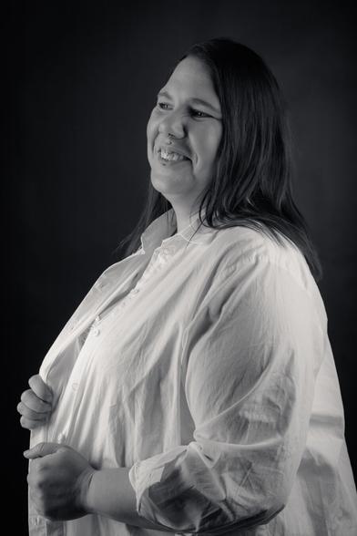 A black-and-white portrait of a woman standing confidently against a dark background. She wears a white button-down shirt over a lace top, with her hands holding the shirt closed. Her long hair falls to one side, and she looks directly at the camera with a calm, self-assured expression. The lighting emphasizes her strong presence and natural beauty.