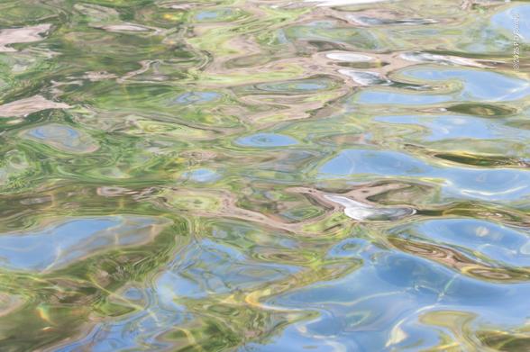 Gentle, glassy rippled patterns in light brown, light green and pale blue; sandstone cliffs, trees and sky reflected in a waterfall plunge pool.