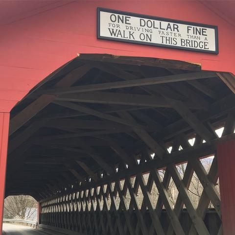 Closeup of the entry into a red covered bridge.  Sign above bridge entry says 'one dollar fine for driving faster than a walk on this bridge'.  Multiple crossing wood support beams can be seen inside, forming diamond and x patterns.  The paved road can be seen at the far end of the bridge, complete with guardrail.