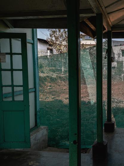 A photo from the inside of an open courtyard in a building. There are three square shaped wooden pillars painted green on the right of the image. To the left of the image, there is a door and a wall, both accented with the same green as the pillar. In the background there is see through green netting, through which a dug out backyard is visible.