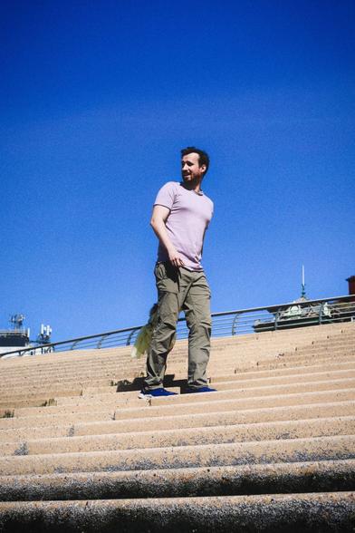 A man wearing a pale purple t-shirt, khaki trousers, and blue shoes stands on wide concrete steps. He is looking to his left and holding a light-colored jacket. Behind him, at the top of the steps, is a curved metal railing. The sky is clear and bright blue.