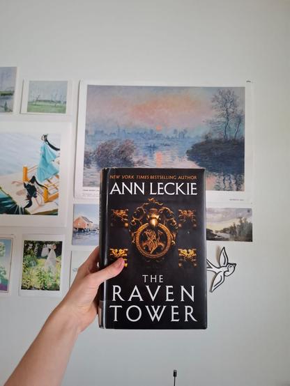 Käsi pitelee Ann Leckien The Raven Tower -kirjaa seinän edessä