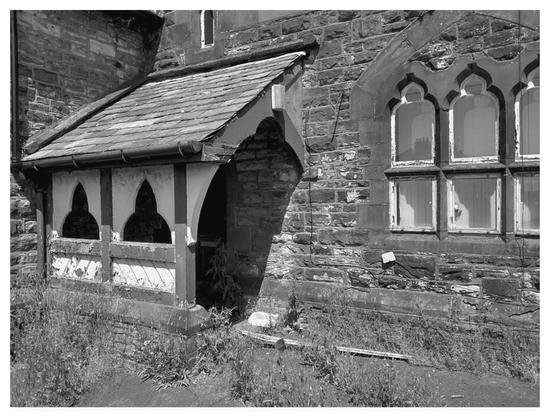 Black and white photograph showing an overgrown porch on an old stone building with elaborately carved windows.