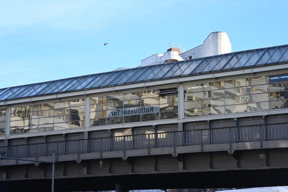 "A view of an elevated train station with a glass facade reflecting distorted images of nearby buildings and a yellow train inside. The sign inside the station, visible in reverse through the glass, reads 'Kottbusser Tor'. A bird flies above the structure against a clear blue sky."

Einschätzung:
Das Bild wirkt urban und lebendig, mit einem Hauch von Berliner Alltag eingefangen durch die bekannte Station „Kottbusser Tor“. Die Spiegelungen in den Glasscheiben geben dem Foto einen interessanten, fast surrealen Touch. Das klare Licht und der Kontrast zwischen dem alten Stahlbau und der modernen Architektur im Hintergrund verstärken den visuellen Reiz
