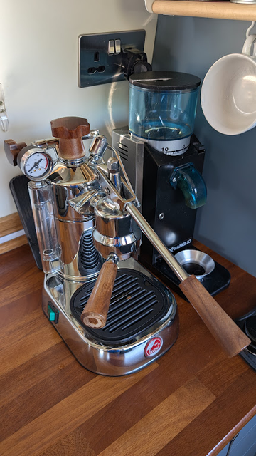 A photo of my La Pavoni PL lever espresso machine, which has after-market brown wooden handles (instead of the standard black bakelite ones). It sits next to the Rancilio Rocky grinder, both on a wooden work surface. There's a white coffee cup hanging on a hook besides the grinder.