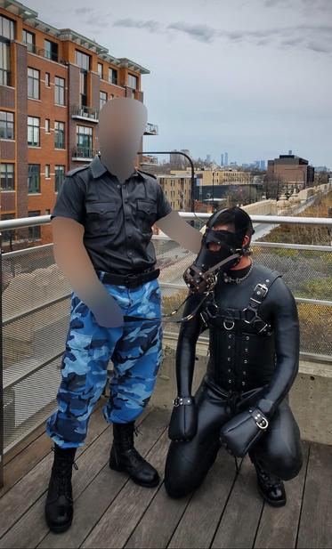 The same pony crouches down while kneeling on right knee. Sir stands to the pony’s right with Their right hand resting on the pony’s neck and Their left hand holding on the leather lead attached to a brown leather muzzle on the pony’s leather snout.