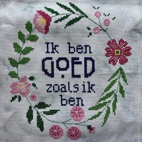 Cross stitch embroidery piece with text in dark blue letters. The text says ‘Ik ben goed zoals ik ben’. Around the text is a wreath of various pink flowers and green leaves.