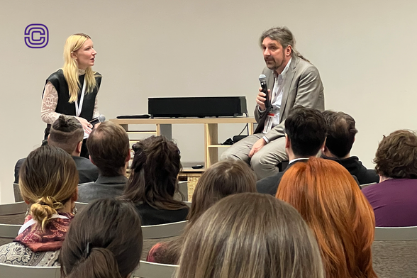 Zwei Personen sitzen auf einer kleinen Bühne vor einem Publikum. Links sitzt eine blonde Frau in schwarzer Weste über einem weißen Spitzenoberteil, sie hält ein Mikrofon locker in der Hand. Rechts sitzt ein Mann, Prof. Dr. Christian Hummert, Forschungsdirektor der Cyberagentur, mit grauem Anzug, langen Haaren und Bart. Er spricht in ein Mikrofon. Zwischen beiden steht ein kleiner Tisch mit einem schwarzen Lautsprecher. Im Vordergrund sind die Rücken und Köpfe zahlreicher Zuhörender zu sehen, die aufmerksam dem Gespräch folgen. Die Szene wirkt konzentriert und offen, die Atmosphäre ist professionell und dialogorientiert. An der Wand links oben ist das Logo der Cyberagentur zu sehen – ein stilisiertes, violettes „C“ in zwei ineinanderliegenden Kreisen. Foto: Cyberagentur