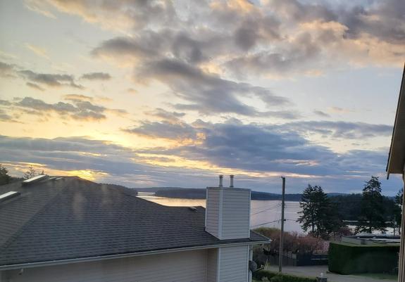 A tranquil scenic view of a sunrise over calm ocean waters. Houses form the foreground, with rooftops and chimneys visible. The landscape transitions into a calm expanse of water, beyond which are forested islands. The sky is filled with clouds streaked with gold.